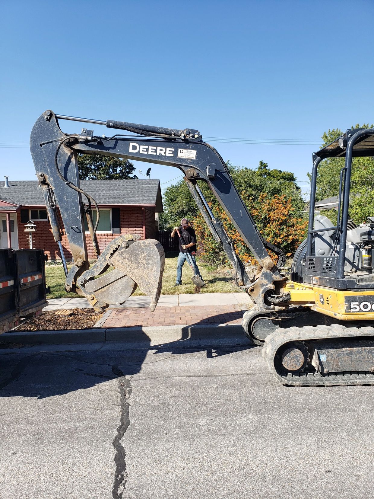 A man shoveling soil near a John Deere excavator on a suburban street.