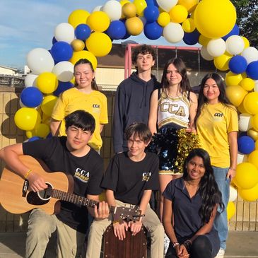 Group of seven young people smiling under a balloon arch, one playing guitar.