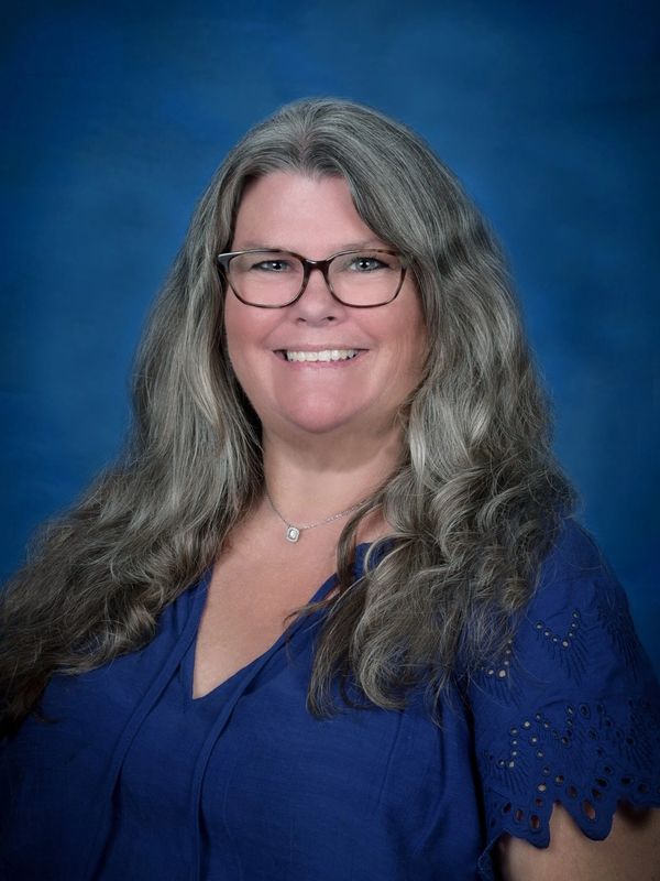 Smiling woman with long gray hair and glasses in a blue blouse.