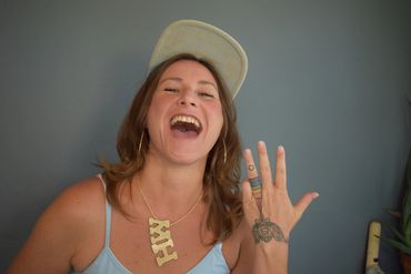 madame harlequin in flat brim hat showing off rainbow hand tattoo with gold MH necklace and hoops