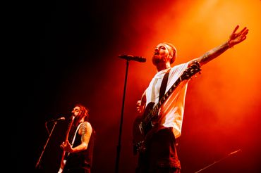 Two tattooed musicians passionately perform on stage under vibrant orange lighting.