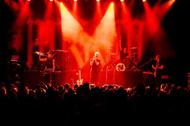A band performs energetically on stage under red lights in front of a large crowd.