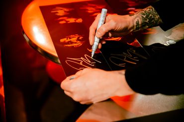 Tattooed hands signing posters with a white marker under warm lighting.