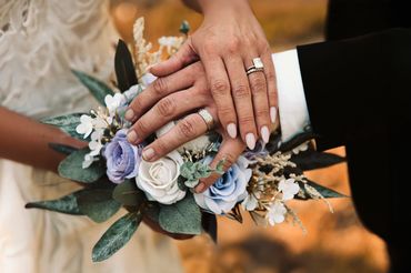 Newlyweds' hands with wedding rings over a bouquet of blue and white flowers.