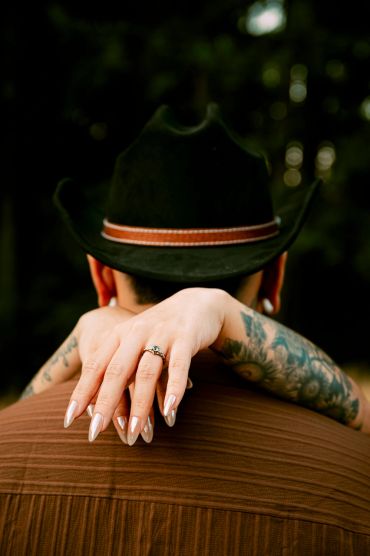 Woman's tattooed arms and engagement ring over man's shoulder in a hat.