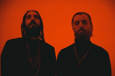 Two men with beards and face tattoos pose against a red background.