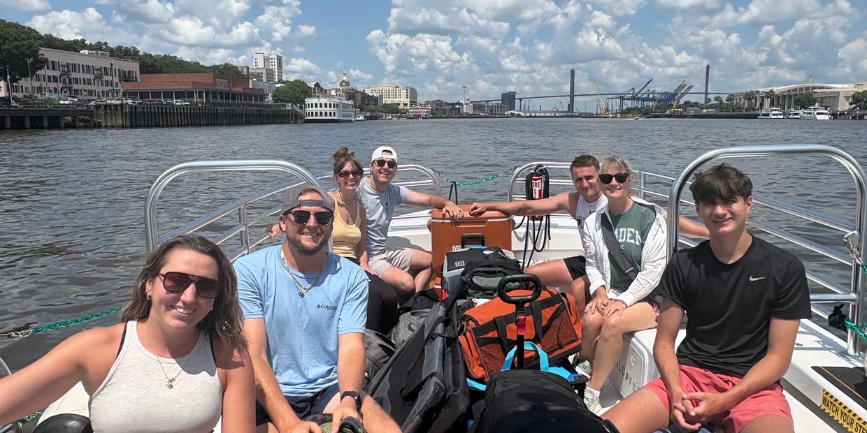 Seven people enjoying a sunny boat ride with city skyline in the background.