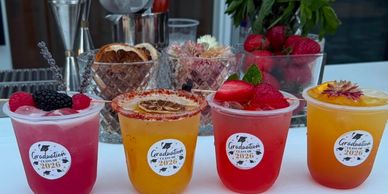 Four colorful graduation-themed drinks with fruit garnishes on a table.