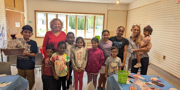 Group of children and two adults smiling in a bright room with craft supplies.