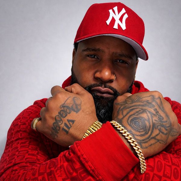 Man in red cap and hoodie showing tattooed fists and gold jewelry.