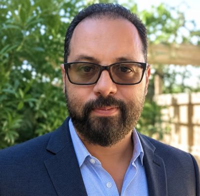 Man wearing glasses and a suit jacket with greenery in background.