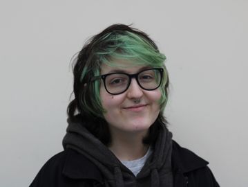 A man with green and black hair and glasses is smiling against a blank background.