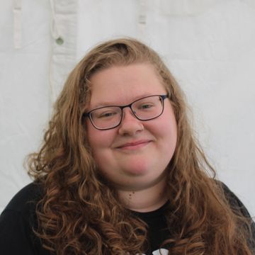 A person smiling against a white background with curly long blond hair wearing glasses.