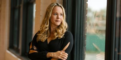 Woman with blonde hair looks thoughtfully out a window, arms crossed.