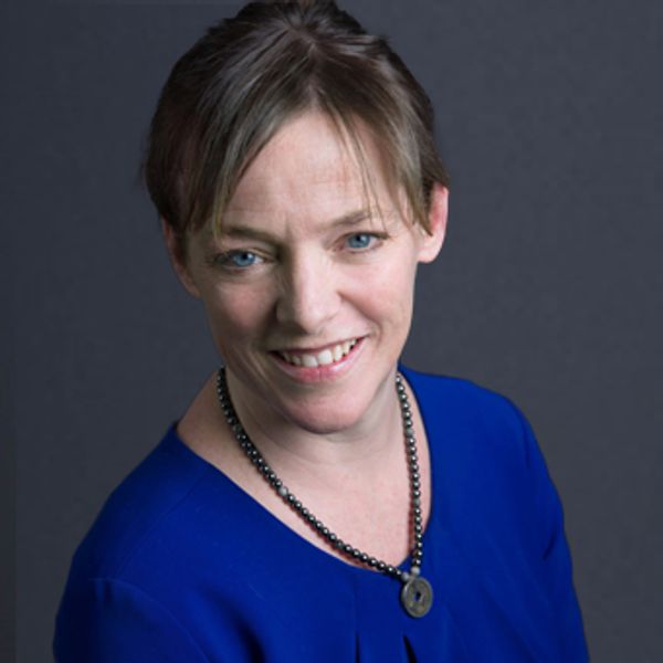 A smiling woman in a blue top with a necklace against a dark background.