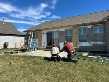 Two workers on grass outside a partially renovated house with ladders and tools.