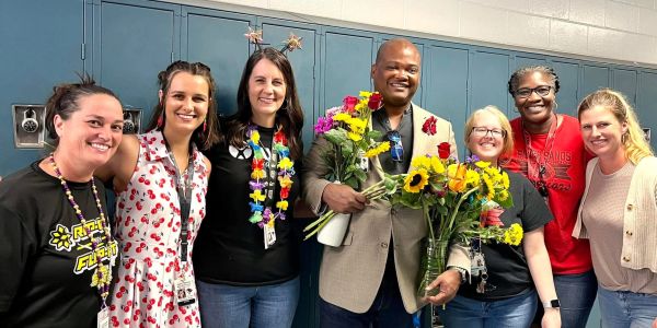 A group of seven smiling people, one holding colorful flowers, standing in front of lockers.