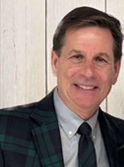 Smiling man in a plaid blazer and tie against a light wood background.