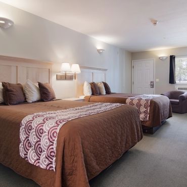 Hotel room with two queen beds, brown bedding, and seating area near window.