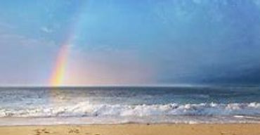 A rainbow appears over the ocean at sunset with gentle waves.