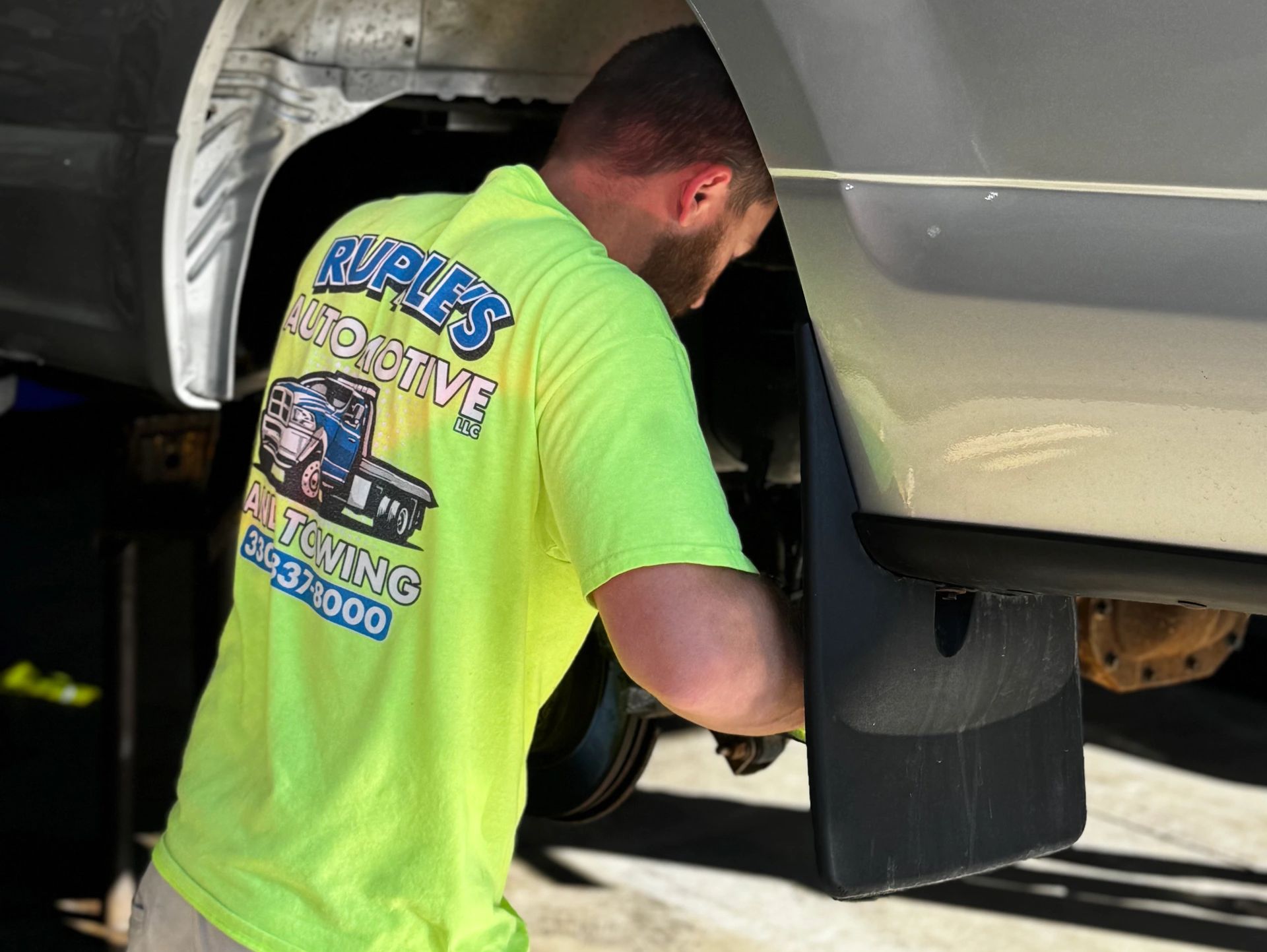 A mechanic in a bright green Ruples Automotive shirt working on a vehicle's wheel area.