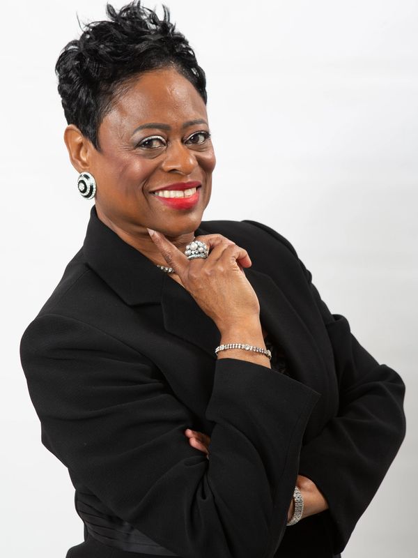 Confident woman in black suit smiling with hand on chin against white background.