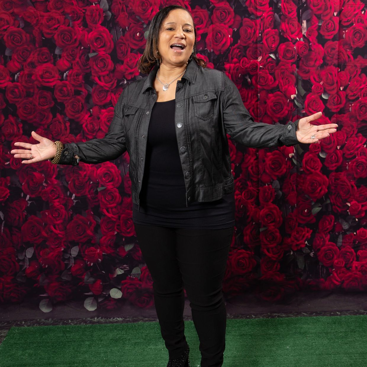 Woman in black outfit stands with arms open against a vibrant red rose backdrop.