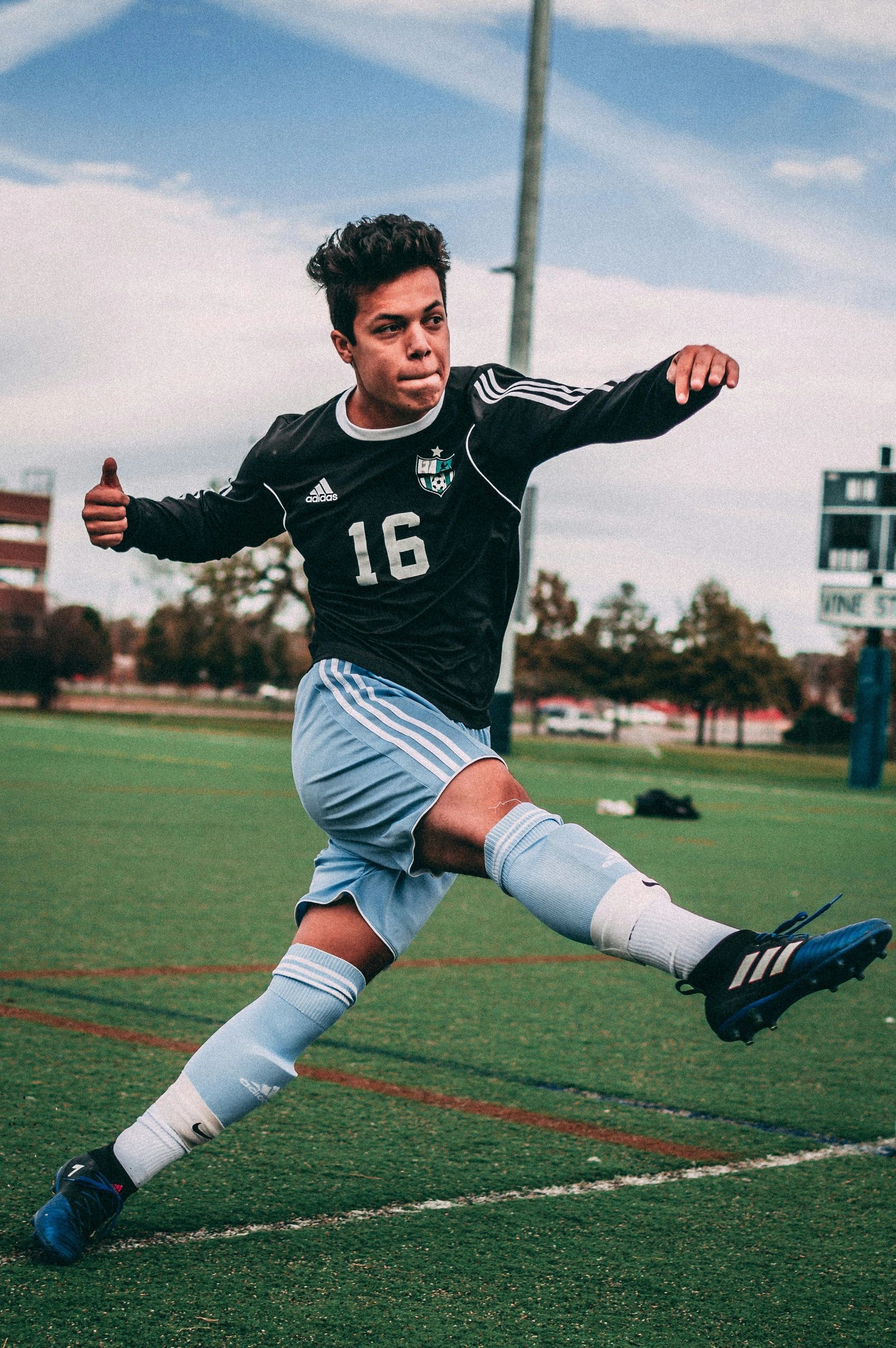 A young person in a jersey mid-kick on a soccer pitch. Green and white background with blue sky.