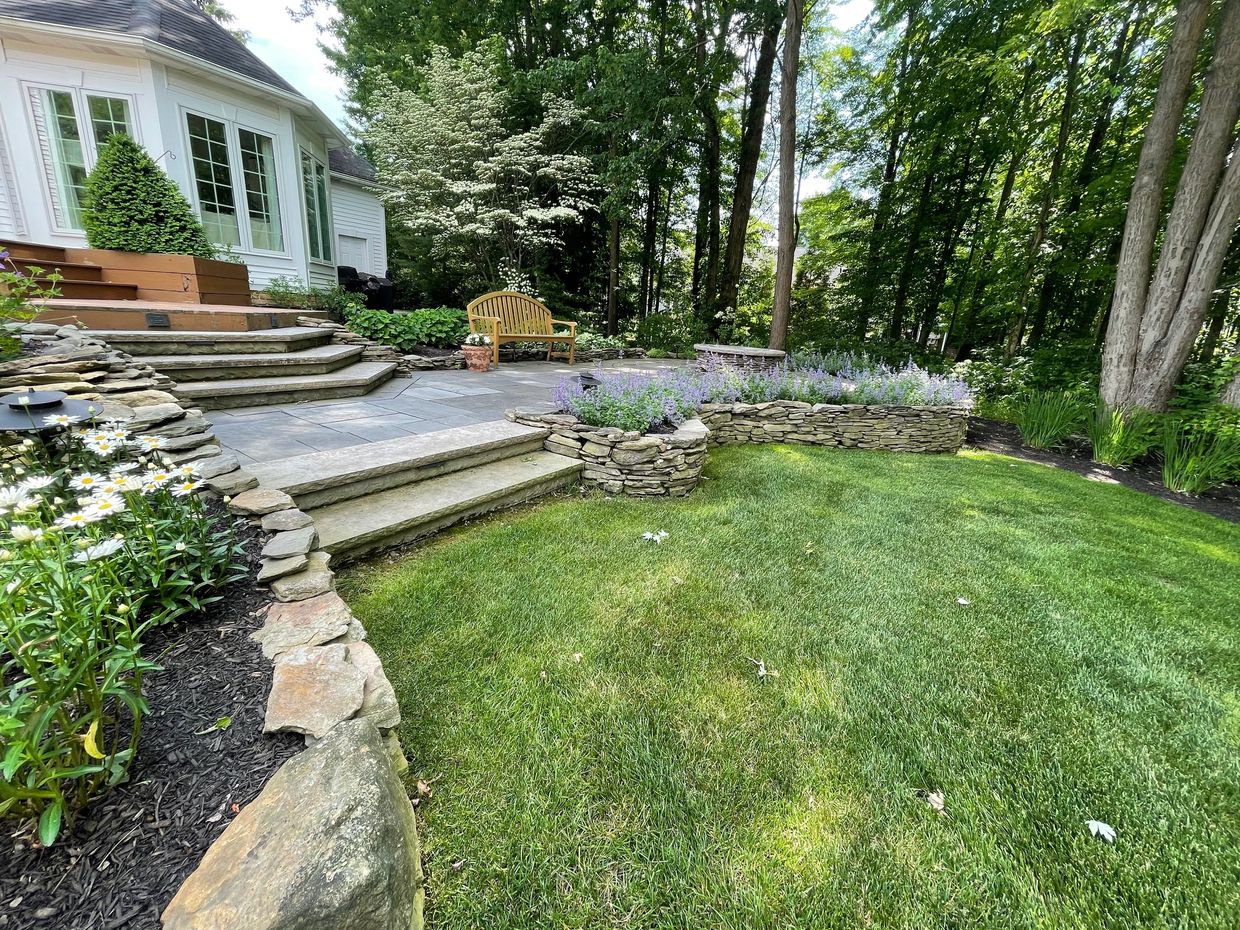 A serene backyard patio with stone steps, flower beds, and a wooden bench surrounded by lush greenery.