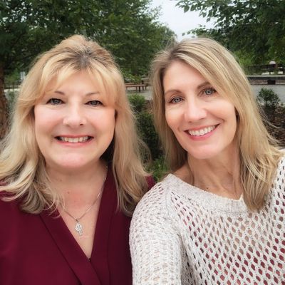 Two smiling women posing outdoors with trees in the background.