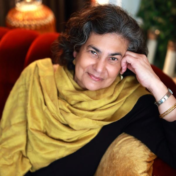 Woman with black and grey hair wearing gold scarf sitting smiling on a red couch