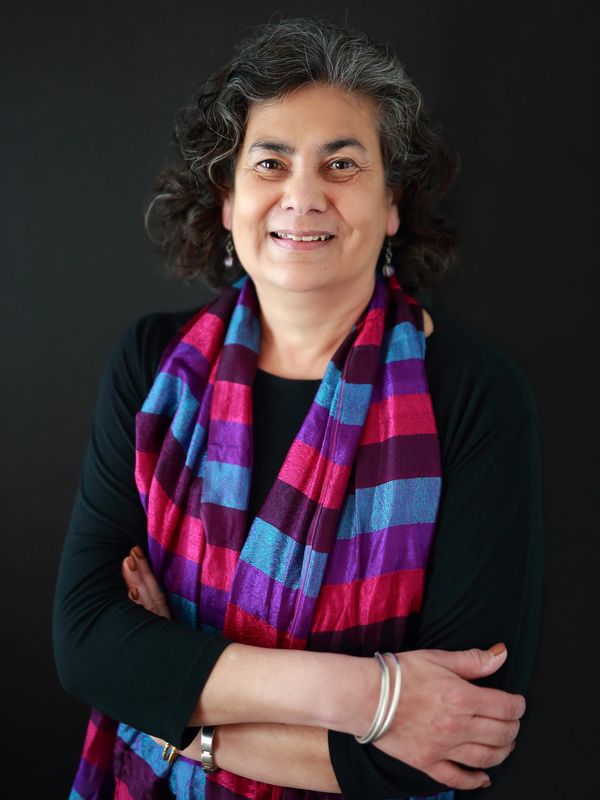 Woman with black hair with grey  wearing purple and blue scarf standing smiling with arms crossed