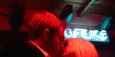 A couple shares a close moment under red light and a neon office sign.