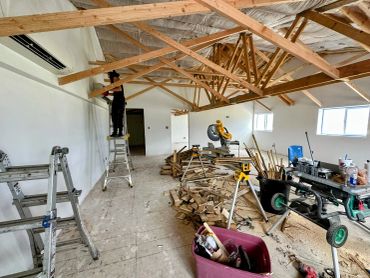 A worker on a ladder installing insulation in a construction site with wood beams and tools scattered.