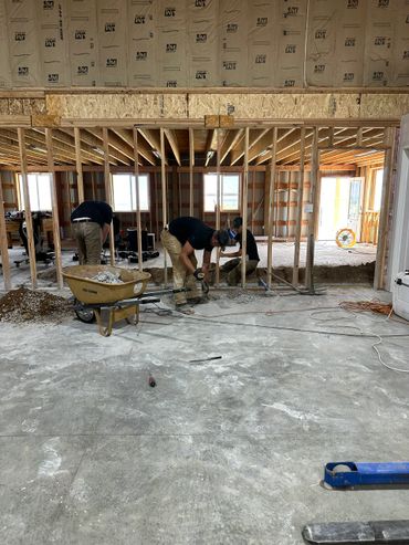 Workers renovating a house interior with exposed framing and concrete floor.