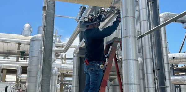 Worker on ladder fixing pipes in an industrial facility on a clear day.