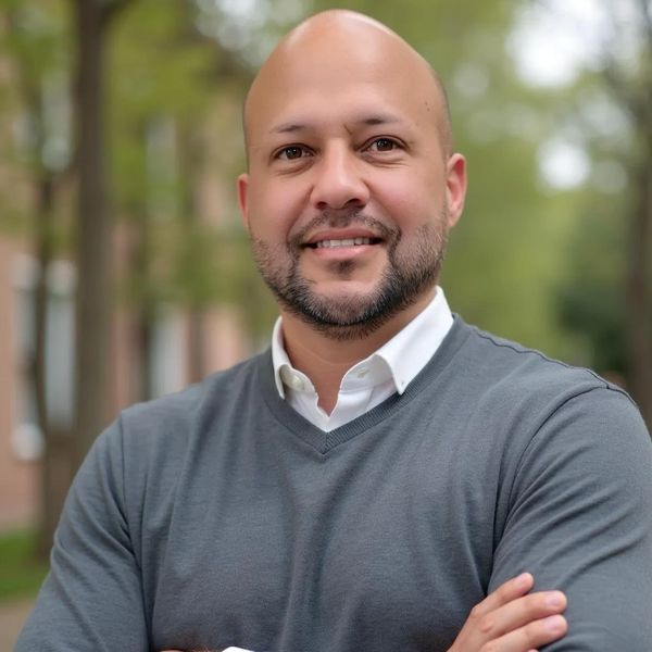 Smiling bald man with a beard in a gray sweater stands confidently outdoors.