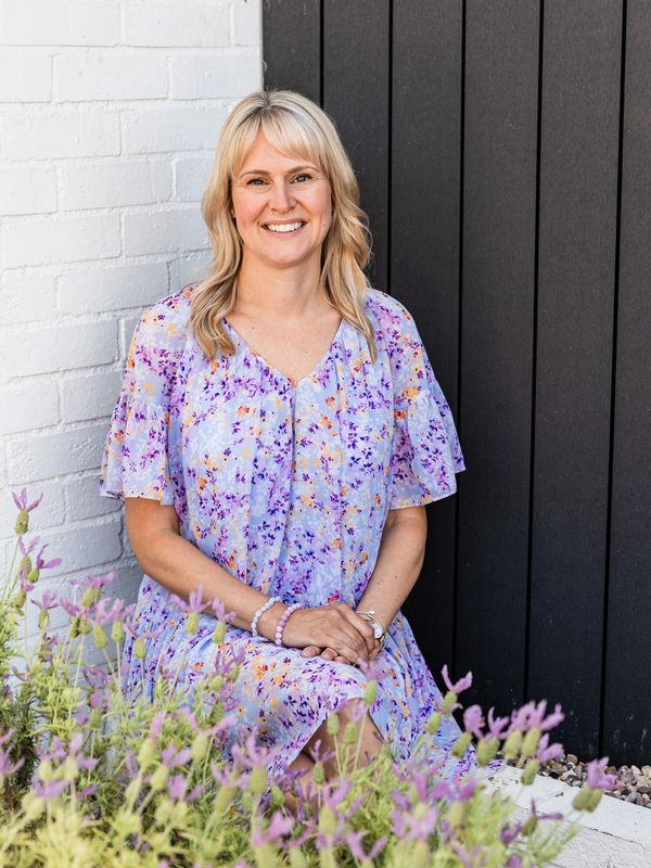 Smiling woman in a floral dress sitting by a garden with purple flowers. This woman is a homeopathy who specialises in natural acne healing and acne scars treatment through being qualified in homeopathy for acne 