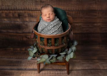 baby wrapped in green blanket, in a brown basket with eucalyptus. brown planks on floor and walls.