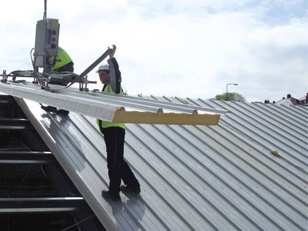 Worker installing insulated metal roofing panel using a suction lifter.