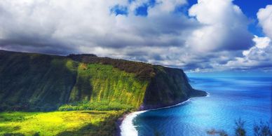 Scenic coastal cliff with lush greenery and bright blue ocean under cloudy sky.