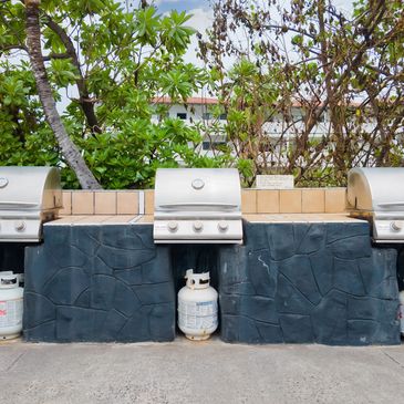 Three outdoor propane gas grills lined up with greenery in the background.