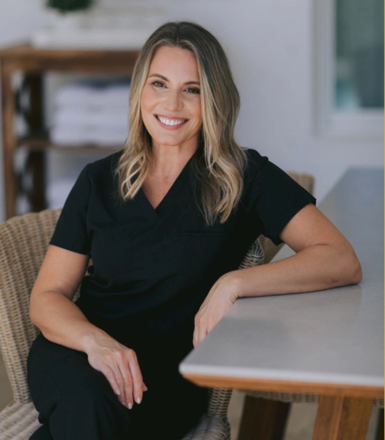Smiling woman in black scrubs sitting on a wicker chair indoors.