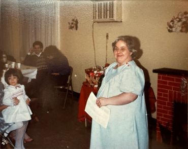 A woman in a light blue dress stands indoors, smiling with people sitting in the background.