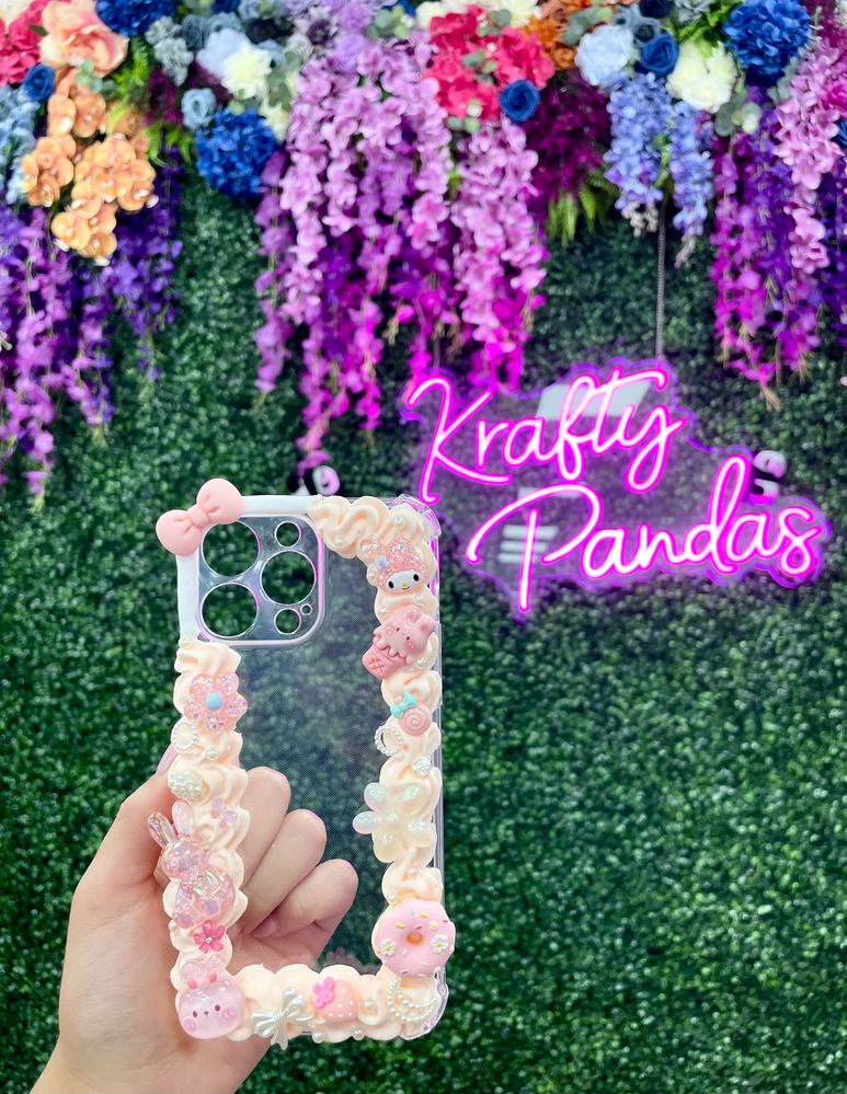 Hand holding a decorated phone case against a floral wall with neon sign.