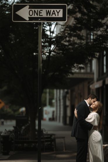 Couple embraces under a one-way street sign in a city setting.