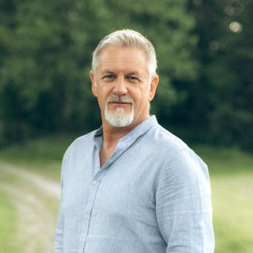 Mature man with gray hair and beard in light blue shirt outdoors.