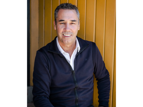 Smiling man in a navy jacket sitting against a yellow wooden wall.