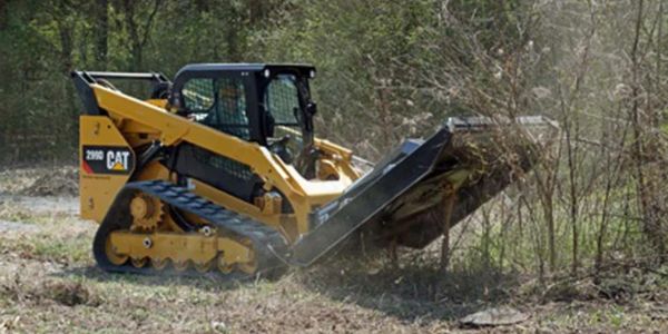 Yellow Caterpillar compact track loader clearing brush in a wooded area.