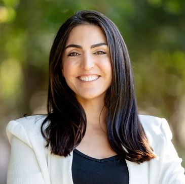Smiling woman with long dark hair wearing a white blazer and black top outdoors.